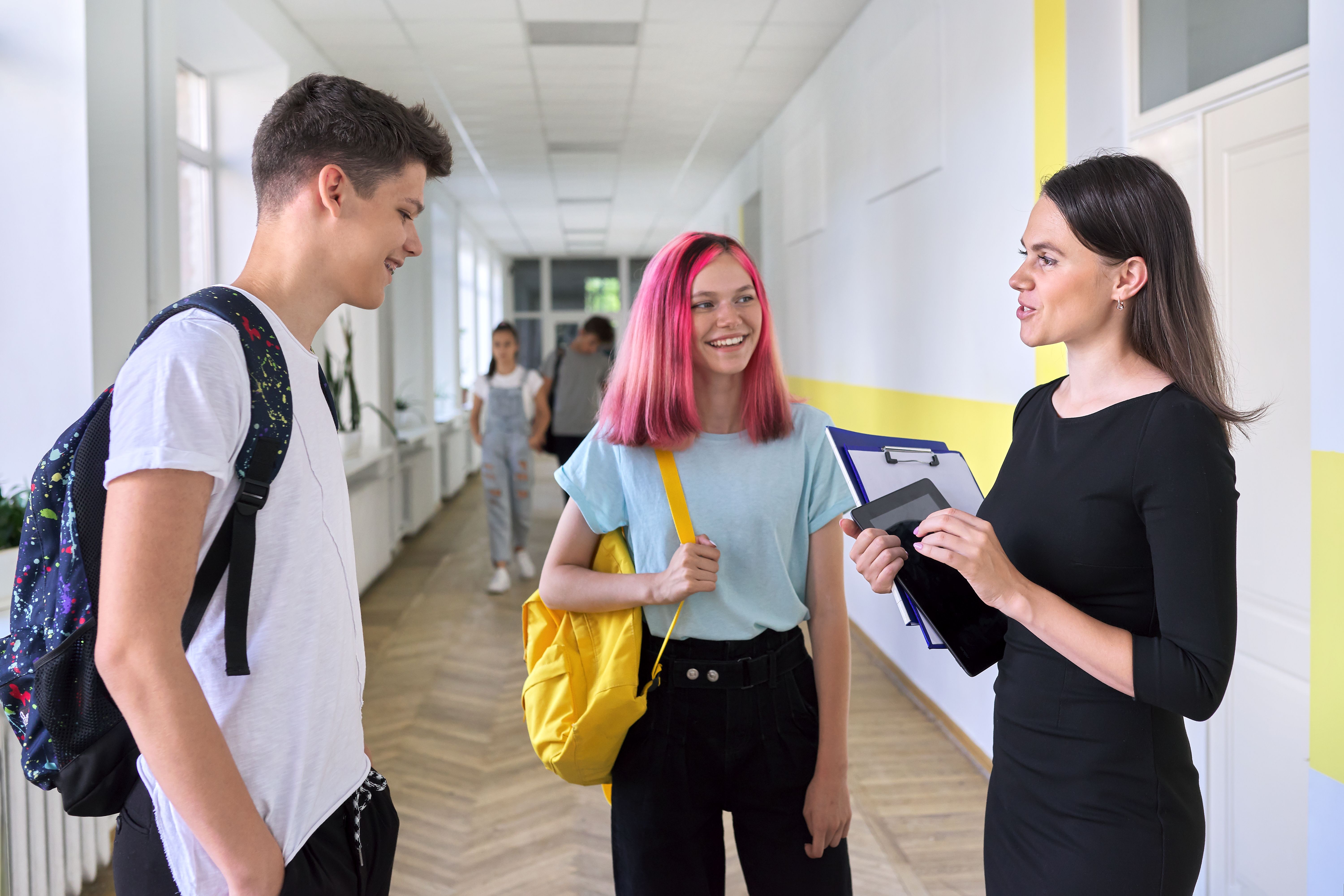 In einem Schulflur stehen eine Schülerin, ein Schüler und eine Lehrerin im entspannten Gespräch zusammen. In einem Schulflur stehen eine Schülerin, ein Schüler und eine Lehrerin im entspannten Gespräch zusammen.