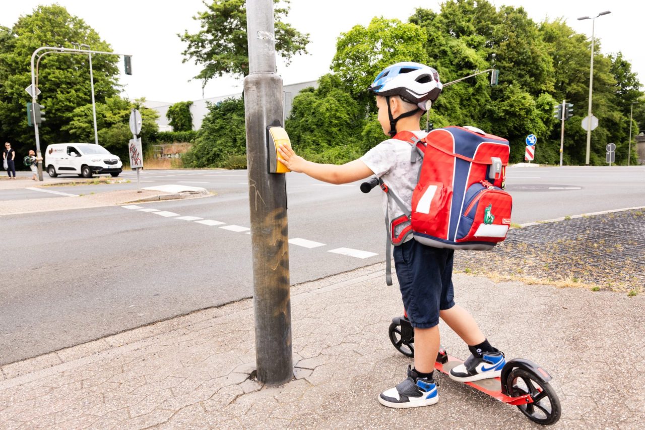 Ein Kind steht mit seinem Roller und einem Schulrucksack auf dem Rücken an einer Ampel.