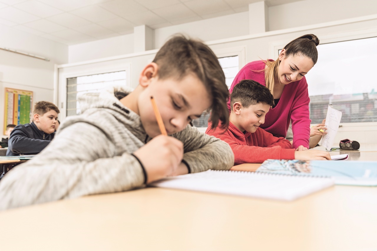 Lehrerin erklärt einem Schüler Unterrichtsstoff in einer Unterrichtssituation. Im Vordergrund sitzt ein anderer, schreibender Schüler. Lehrerin erklärt einem Schüler Unterrichtsstoff in einer Unterrichtssituation. Im Vordergrund sitzt ein anderer, schreibender Schüler.