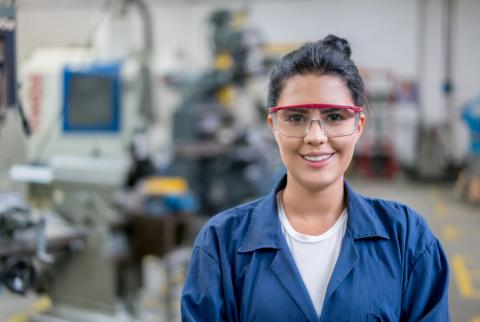 Junge Frau in Arbeitskleidung und Schutzbrille, im Hintergrund Maschinen.