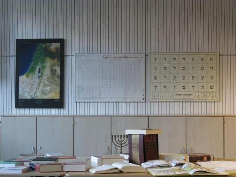 Hebräisch-Klassenraum mit Karte Israels, Schriftzeichen-Plakat, aufgeschlagenen Büchern und siebenarmigem Leuchter.