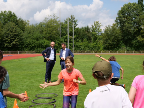Kinder trainieren auf dem Sportplatz. Im Hintergrund schaut Staatssekretär Dr. Mauer ihnen dabei zu.