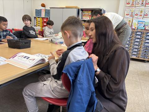 Lehrerin hockt neben einem an einem Tisch sitzenden Kind und erklärt etwas in einem Buch.