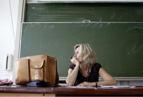 Lehrerin sitzt nachdenklich am Tisch vor einer Tafel.