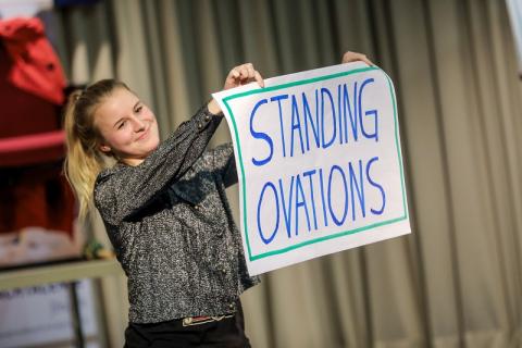 Jugendliche hält in handgeschriebenes Plakat mit dem Schriftzug "Standing Ovations" lächelnd hoch. 