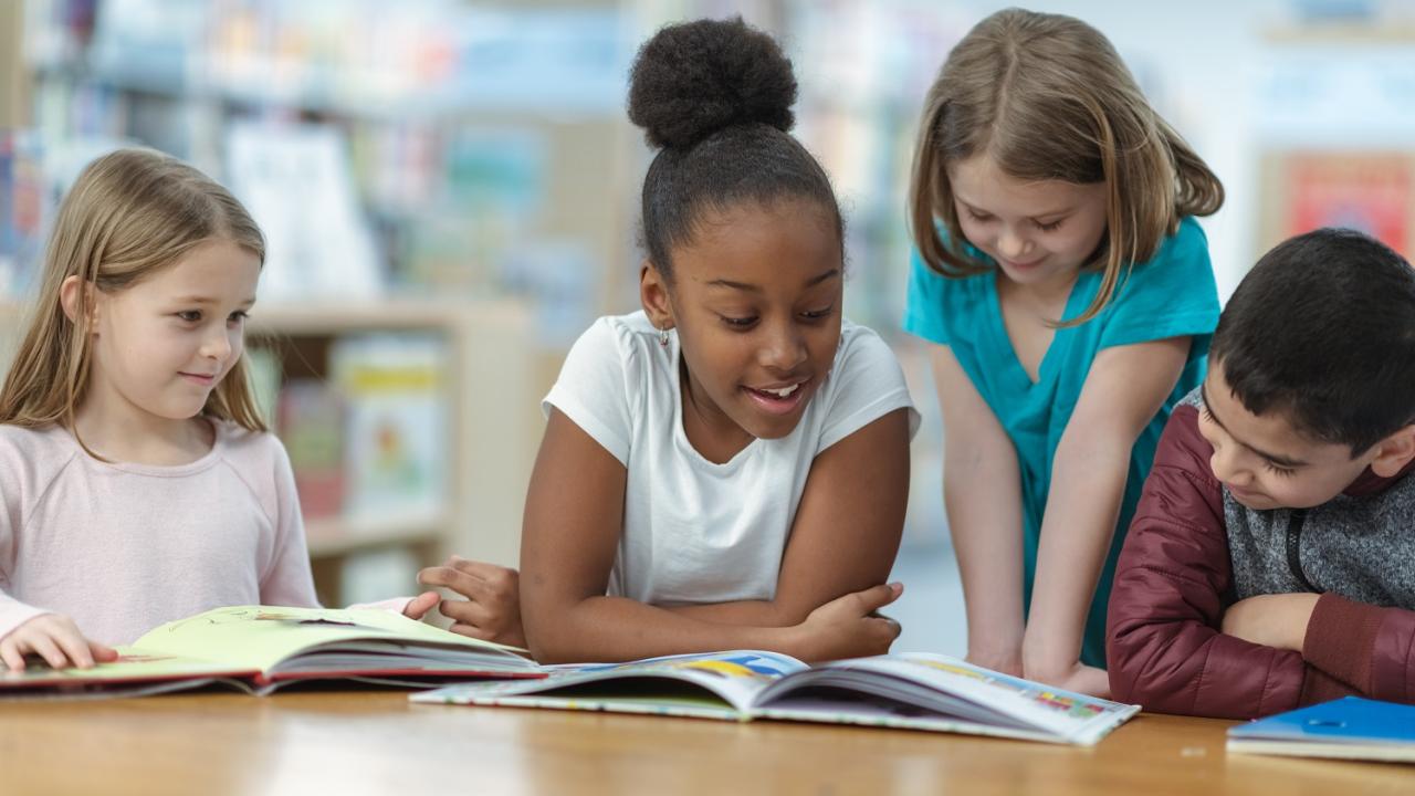 Grundschülerinnen und Grundschüler sitzen an einem Tisch vor aufgeschlagenen Büchern, ein Mädchen liest vor und die anderen hören zu.