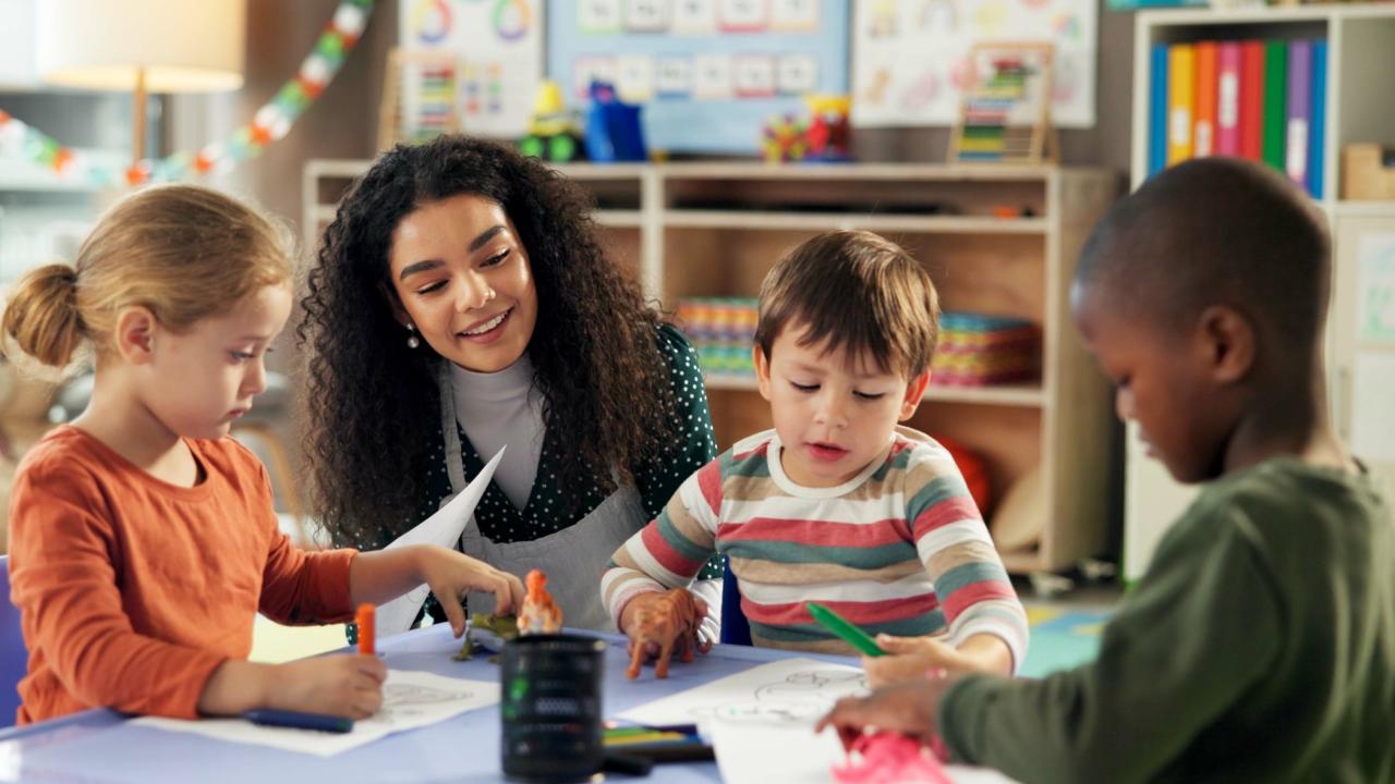 Symbolbild: Lehrerin spricht mit einer Gruppe Kinder, die an einem Tisch sitzen.