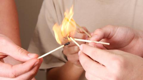 Mehrere Hände halten brennende Streichhölzer zusammen, zu erkennen ist in der Mitte nur ein Kind, das ebenfalls ein Streichholz in die große Flamme streckt. 