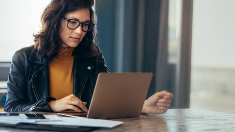 Eine Frau sitzt an einem Schreibtisch und schaut in einen aufgeklappten Laptop.