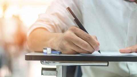 Detailaufnahme einer Hand, die auf Papier schreibt. Im Hintergrund unscharf weitere schreibende Personen an Tischen.