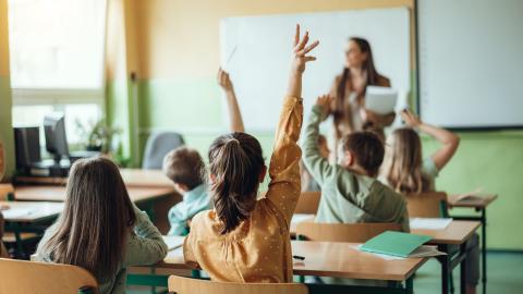 Rückansicht von Grundschülerinnen und Grundschülern, die in einem Klassenraum an ihren Tischen sitzen. Im Hintergrund ist unscharf eine Lehrerin zu erkennen.