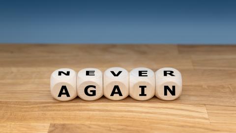 Auf einer Holzplatte liegen fünf Buchstabenwürfel, die die englischen Worte "NEVER" und "AGAIN" bilden.