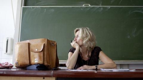 Lehrerin sitzt nachdenklich am Tisch vor einer Tafel.