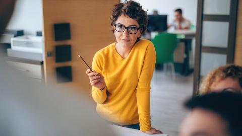 Frau setzt zum Sprechen an, dabei stützt sie sich auf einen Tisch auf. Ihre Gesprächspartner sind nur von Hinten zu erkennen. 