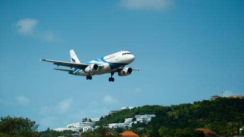 Blauer Himmel, davor startendes oder landendes Flugzeug, im Hintergrund bewaldete Landschaft mit weißen Häusern.