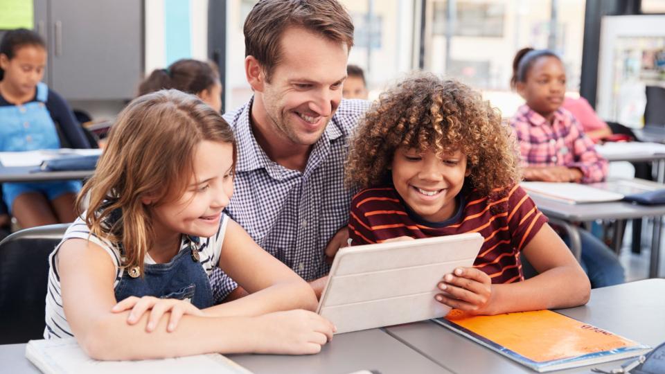 Lehrer, eine Schülerin und ein Schüler blicken in einem Klassenraum gemeinsam auf ein Tablet (Computer). 