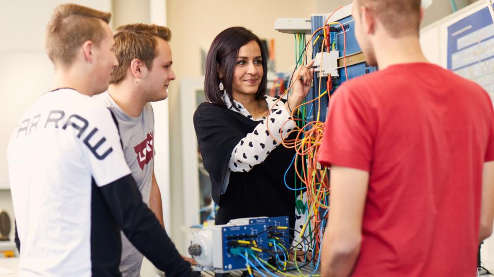 Eine Lehrerin erklärt jugendlichen eine elektrische Apparatur.