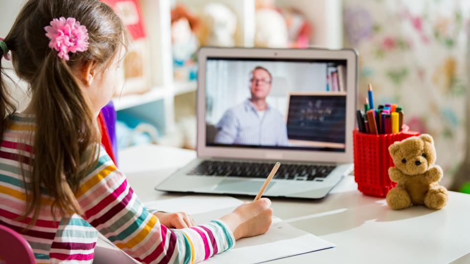 Eine Schülerin blickt beim Lernen auf Distanz auf ihren Laptop. Auf dem Bildschirm des Laptops ist verschwommen ein Lehrer mit einer Tafel neben sich abgebildet