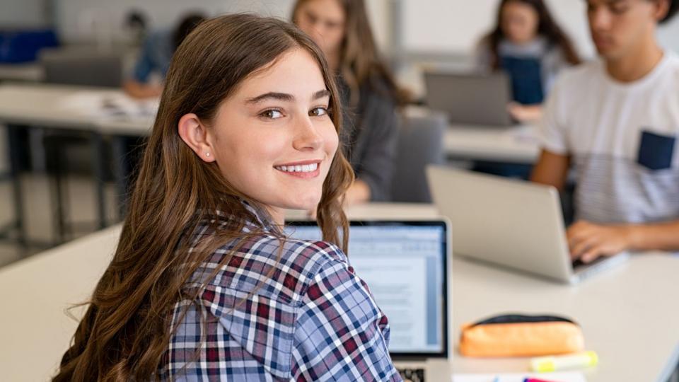 Eine Schülerin sitzt in einer Unterrichtssituation am Laptop und wendet sich dem Betrachter zu. Eine Schülerin sitzt in einer Unterrichtssituation am Laptop und wendet sich dem Betrachter zu.