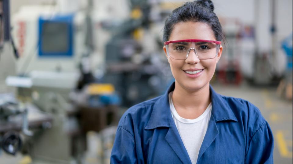 Junge Frau in Arbeitskleidung und Schutzbrille, im Hintergrund Maschinen.