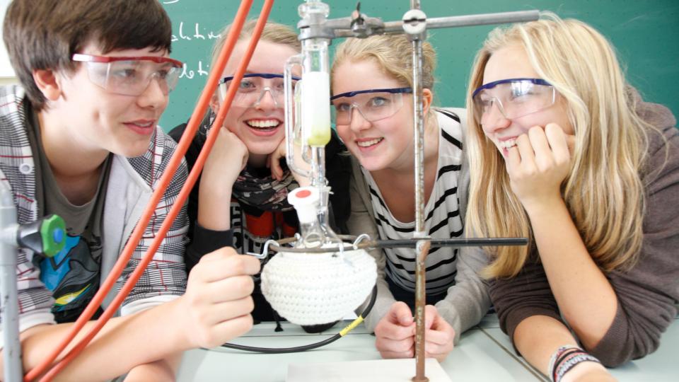 Drei Schülerinnen und ein Schüler betrachten einen chemischen Versuchsaufbau.