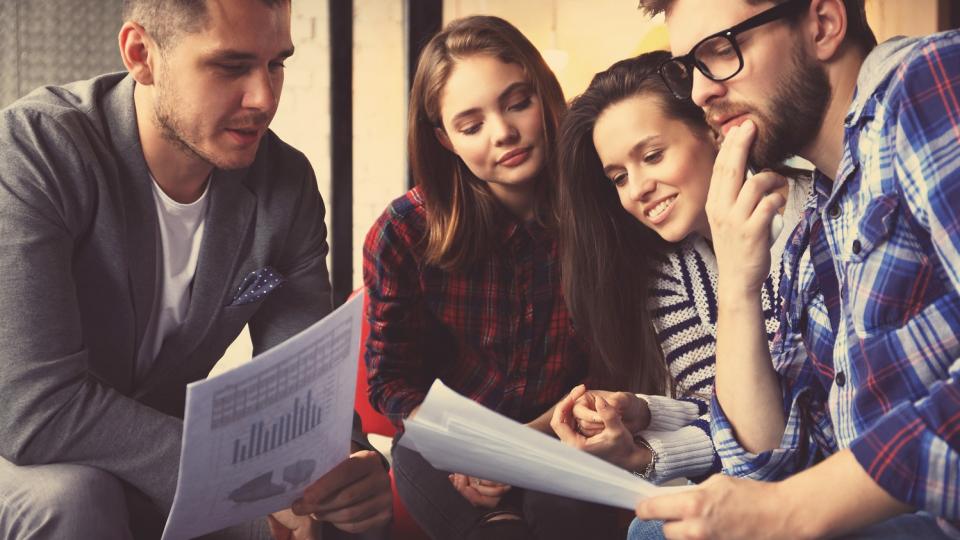 Eine Gruppe von Schülerinnen und Schülern sitzt zusammen und spricht über Wirtschaftsdokumente, die zwei von ihnen in den Händen halten. Eine Gruppe von Schülerinnen und Schülern sitzt zusammen und spricht über Wirtschaftsdokumente, die zwei von ihnen in den Händen halten.