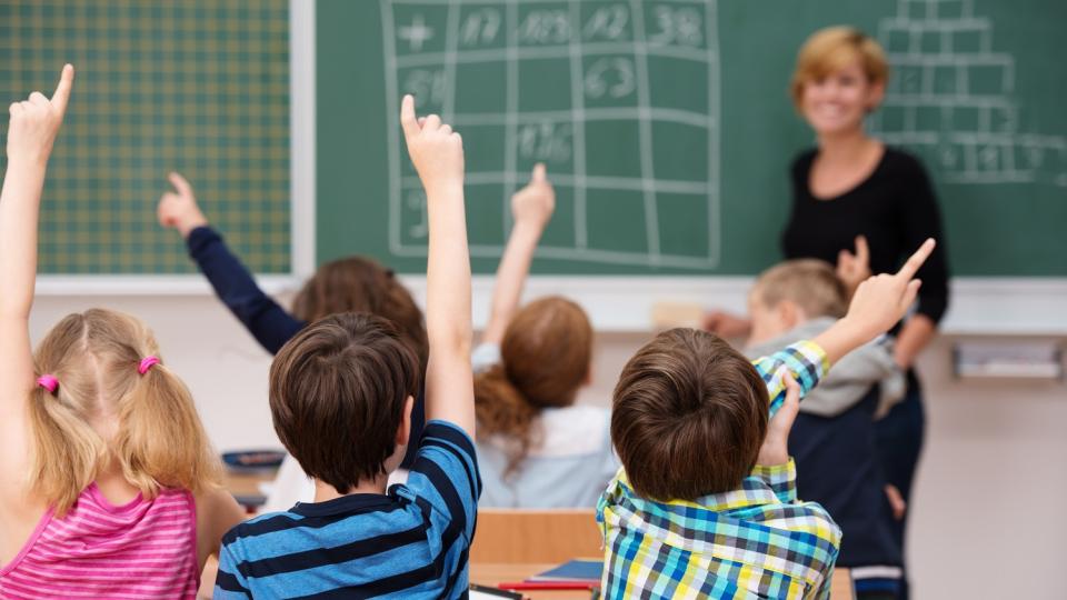 Grundschülerinnen und -schüler melden sich, im Hintergrund eine Tafel mit Rechenaufgaben