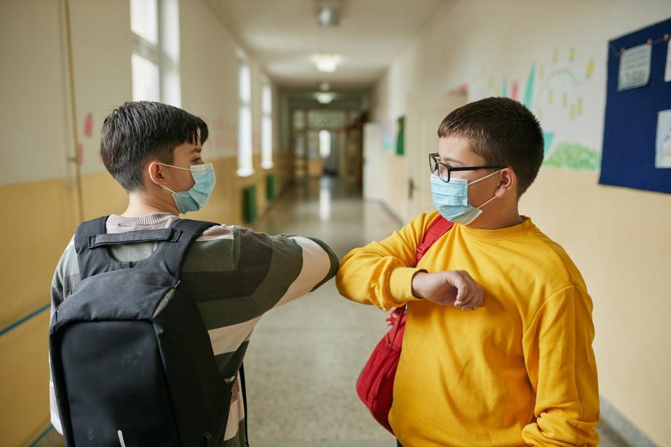 Zwei Alltagsmasken tragende Schüler begrüßen sich mit einem Ellenbogencheck auf einem Schulflur.