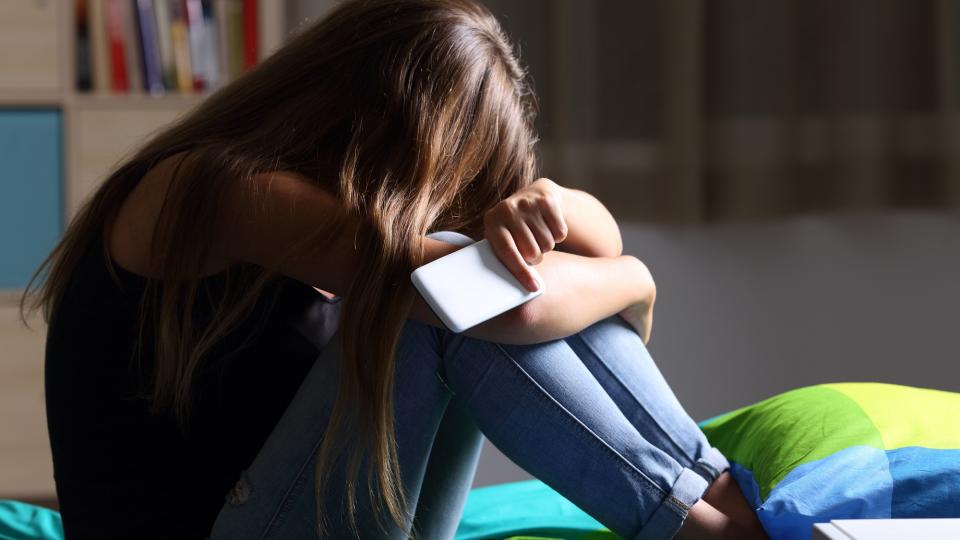 Mädchen hockt auf einem Bett, den Kopf auf die Knie gestützt, in der Hand ein Smartphone.