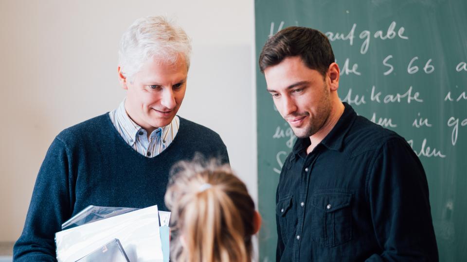 Ein älterer und ein jüngerer Lehrer unterhalten sich mit einer Schülerin.