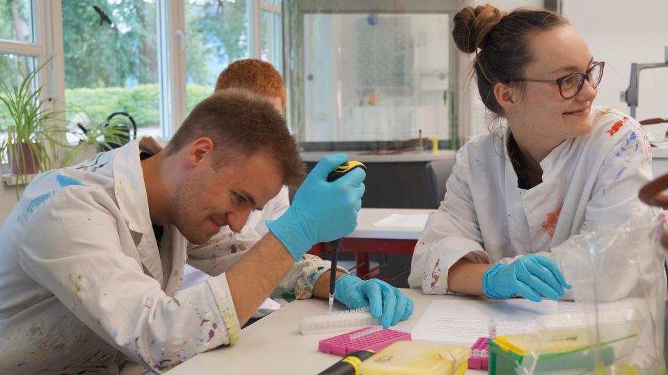 Ein Schüler und eine Schülerin in farbbeklecksten Kitteln führen Versuche mit Pipetten durch Ein Schüler und eine Schülerin in farbbeklecksten Kitteln führen Versuche mit Pipetten durch