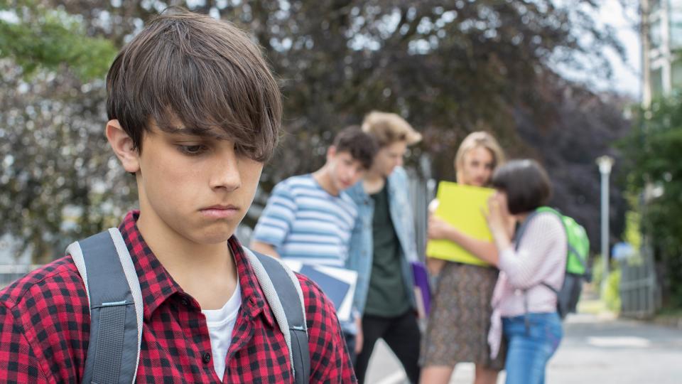 Ein Schüler steht abseits von Gleichaltrigen im Vordergrund, die anderen Jugendlichen scheinen über ihn zu sprechen. Ein Schüler steht abseits von Gleichaltrigen im Vordergrund, die anderen Jugendlichen scheinen über ihn zu sprechen.
