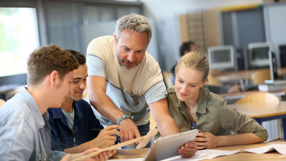 Ein Gruppe älterer Schüler blickt gemeinsam mit einem Lehrer auf ein Tablet Ein Gruppe älterer Schüler blickt gemeinsam mit einem Lehrer auf ein Tablet