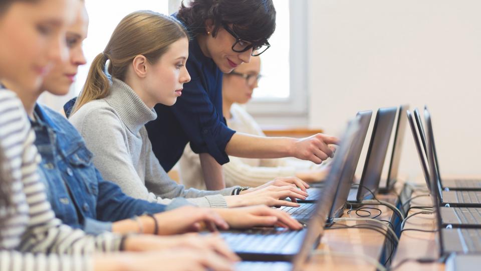 Schülerinnen und Lehrkraft arbeiten jeweils an einem Laptop.