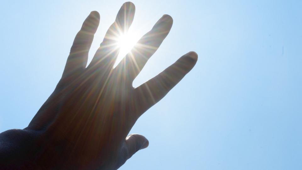 Eine linke Hand vor blauem Himmel, durch die Finger blinzelt die Sonne hindurch.