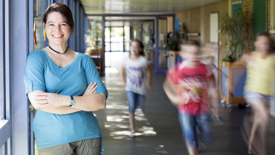 Lehrerin steht lächelnd in einem Schulflur, unscharf  im Hintergrund zu erkennen sind laufende Schülerinnen und Schüler.