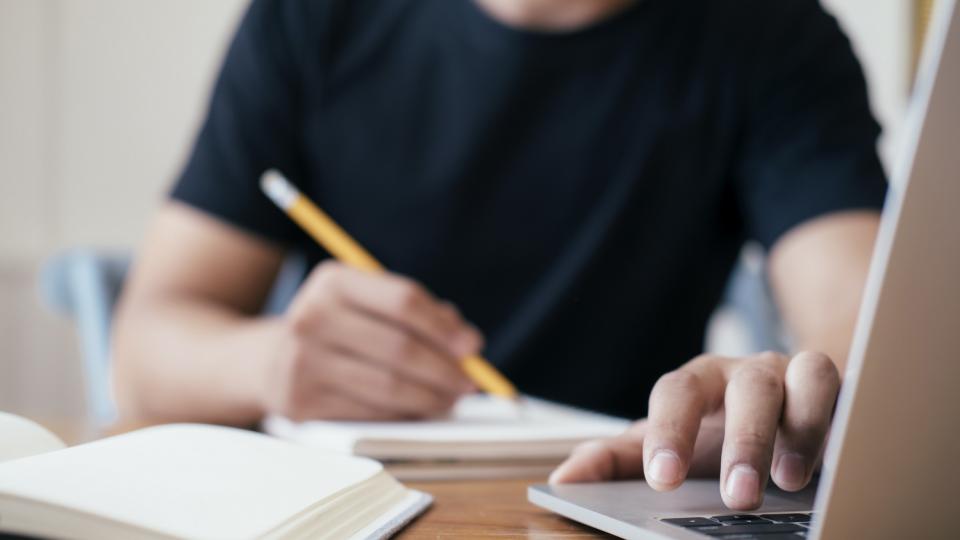 Eine Person, deren Gesicht nicht zu erkennen ist, sitzt an einem Schreibtisch, mit der einen Hand schreibend, die andere Hand liegt auf der Tastatur eines Laptops.