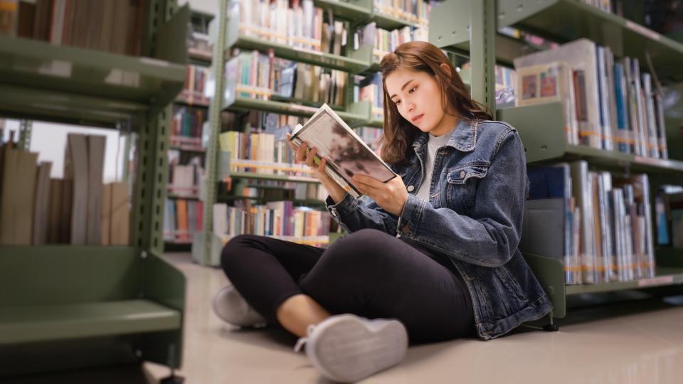 Eine Jugendliche sitzt lesend an ein Regal gelehnt in einer Bibliothek. Eine Jugendliche sitzt lesend an ein Regal gelehnt in einer Bibliothek.