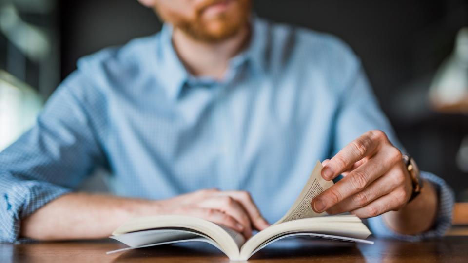 Nahaufnahme: Auf einem Tisch liegt ein geöfffnetes Buch, eine Hand hält es offen, die andere blättert darin. Im Hintergrund ist unscharf die lesende Person zu erkennen.