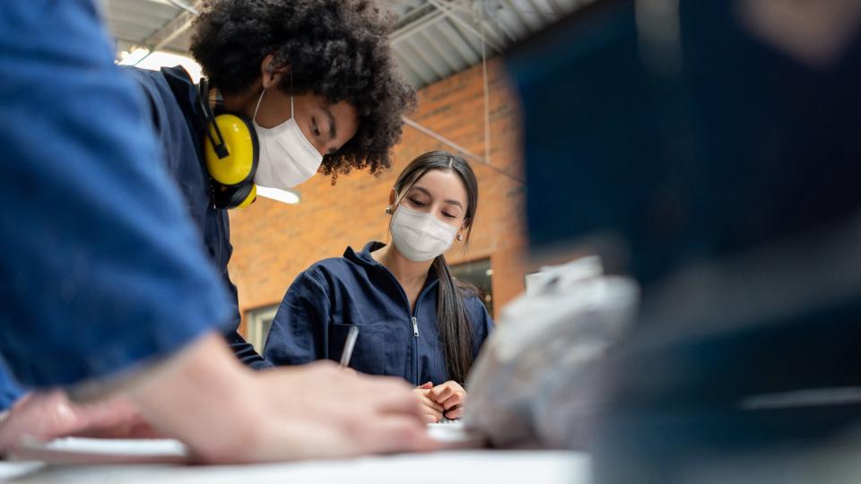 Jugendliche in Arbeitskleidung arbeiten gemeinsam über einen Tisch gebeugt. 