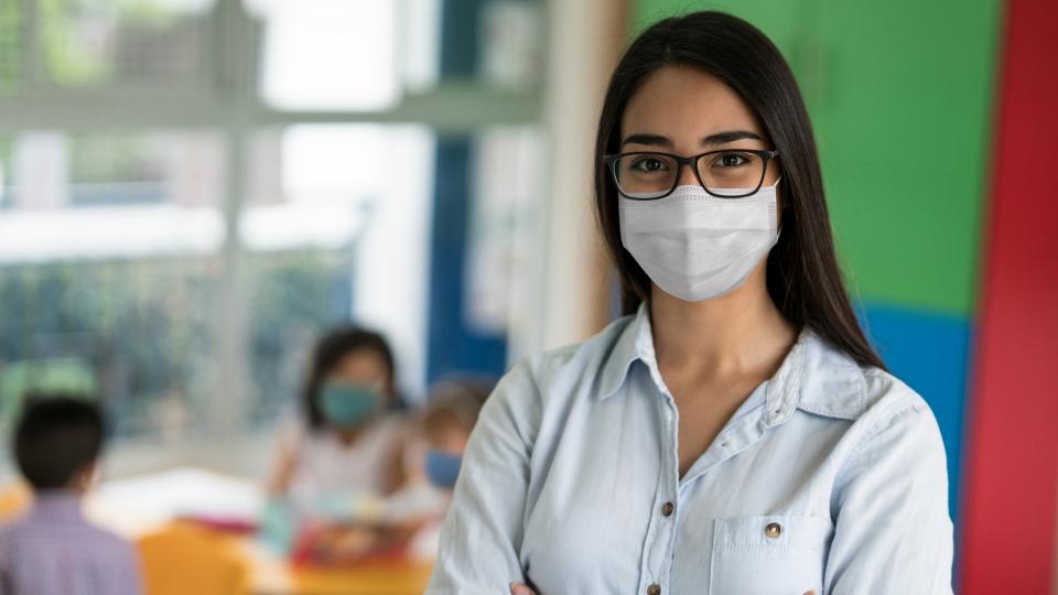 Lehrerin in einem Klassenraum, sie trägt einen medizinischen Mund-Nasen-Schutz.