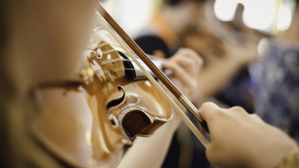 Nahaufnahme einer Geige spielenden Person, das Gesicht liegt in der Unschärfe. Ebenfalls unscharf im Hintergrund sind weitere Musiker/innen zu erkennen. Nahaufnahme einer Geige spielenden Person, das Gesicht liegt in der Unschärfe. Ebenfalls unscharf im Hintergrund sind weitere Musiker/innen zu erkennen.