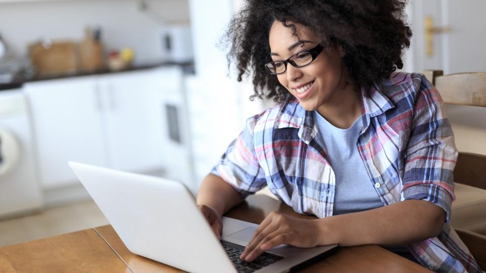 Eine Frau sitzt an einem Tisch und blickt in einen geöffneten Laptop.