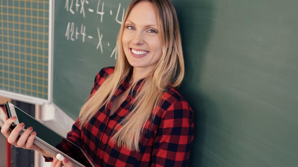 Eine Lehrerin steht mit dem Rücken zur Tafel, in ihren Händen hält sie einen Tablet-Computer.