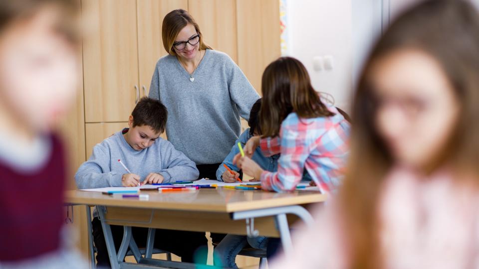 Unterrichtssituation in einer Grundschule: Schülerinnen und Schüler arbeiten, eine Lehrerin blickt ihnen über die Schulter. 