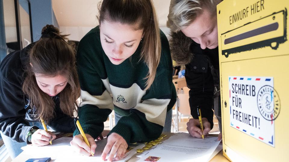 Drei Schülerinnen und Schüler schreiben im Rahmen des Amnesty International-Briefmarathons einen Brief. Drei Schülerinnen und Schüler schreiben im Rahmen des Amnesty International-Briefmarathons einen Brief.