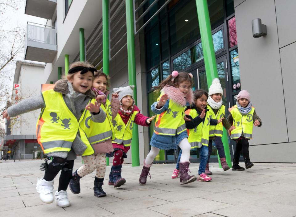 Mehrere Kinder im Kita-Alter laufen in Sicherheitswesten vor einem Gebäude nebeneinander her Mehrere Kinder im Kita-Alter laufen in Sicherheitswesten vor einem Gebäude nebeneinander her