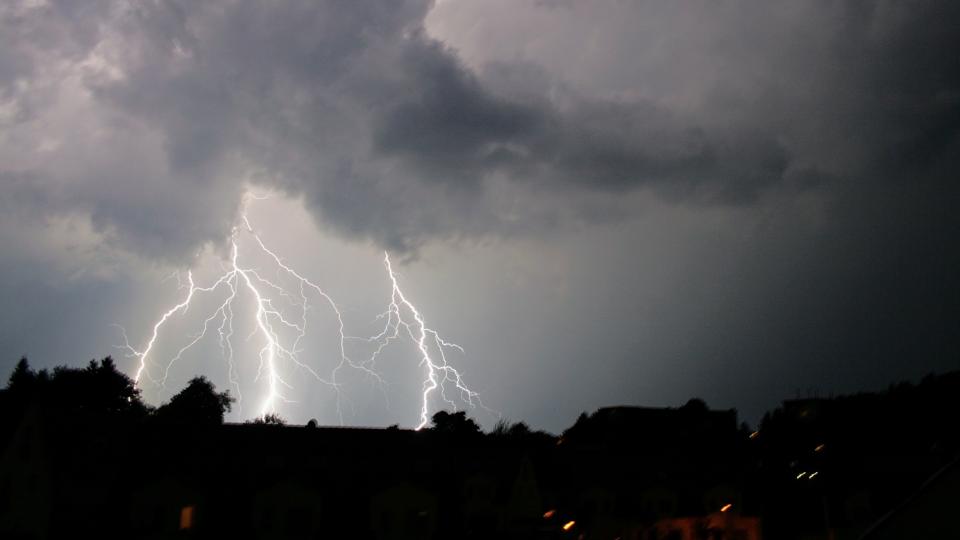Wolkenverhangener Himmel über einer dunkel zu erkennenden Stadt. Aus den Wolken schlagen mehrere Blitze nieder.