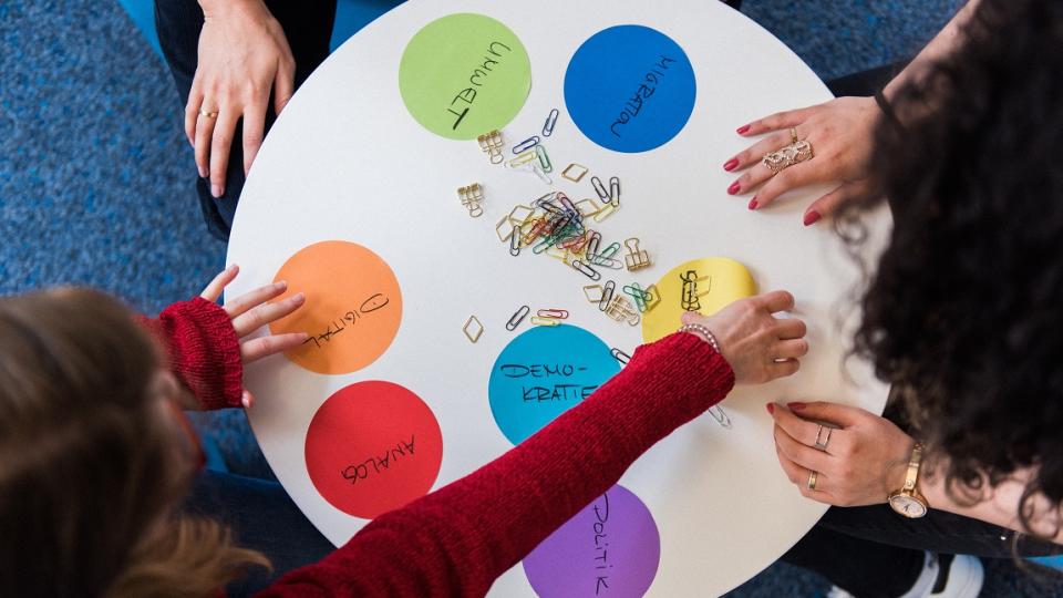 Kinder und Jugendliche sitzen um einen runden Tisch und arbeiten mit Moderationskarten mit Begriffen aus dem gesellschaftspolitischen Bereich. Kinder und Jugendliche sitzen um einen runden Tisch und arbeiten mit Moderationskarten mit Begriffen aus dem gesellschaftspolitischen Bereich.