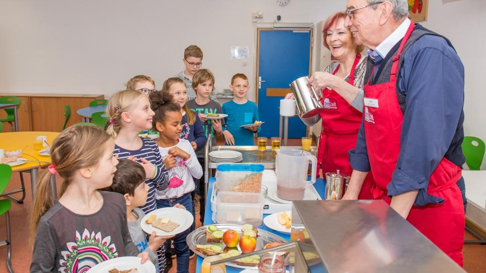 Kinder stehen vor einem Tresen, an dem zwei Erwachsene Frühstück ausgeben. Kinder stehen vor einem Tresen, an dem zwei Erwachsene Frühstück ausgeben.